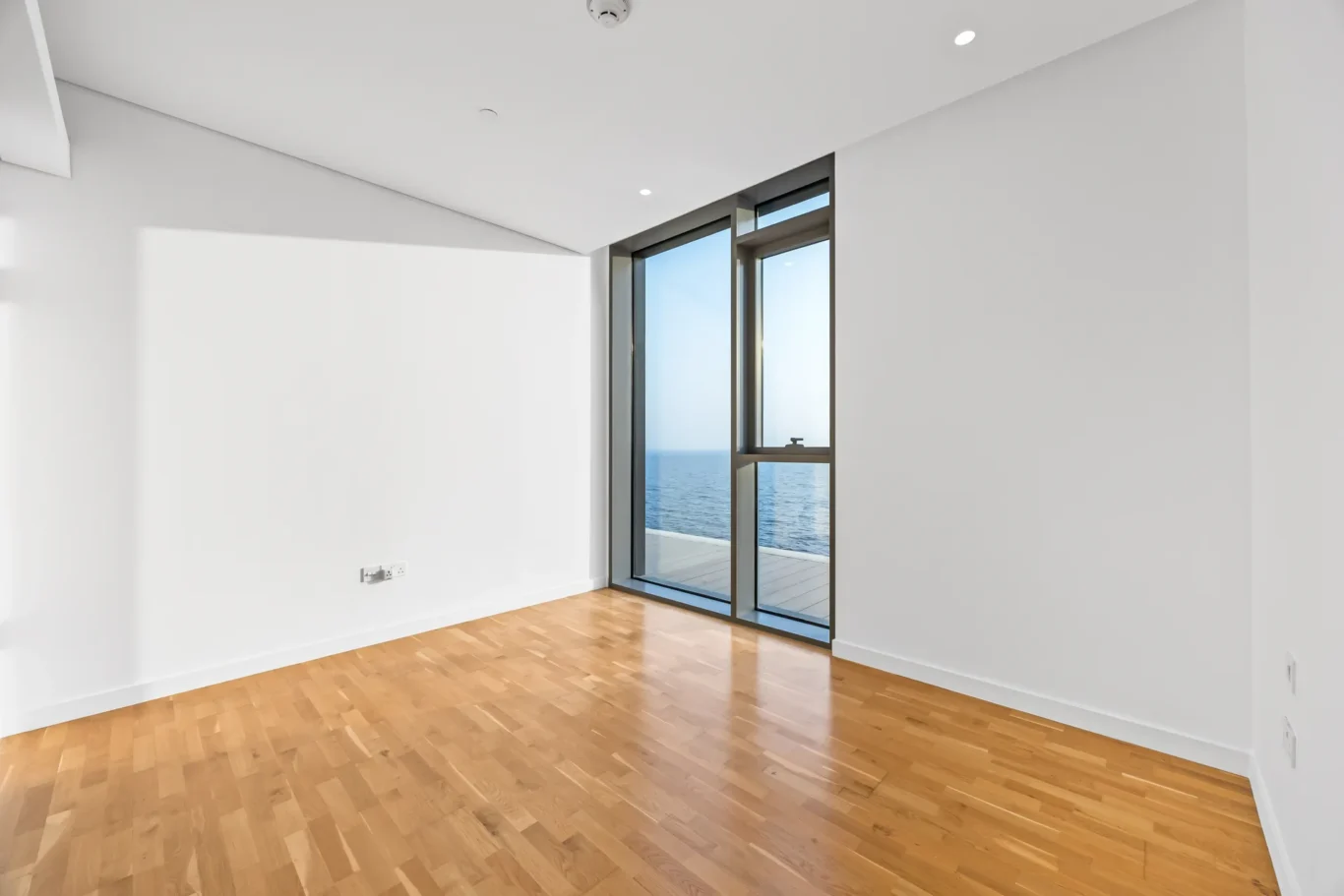 Bright empty room with white walls, light hardwood floor, and large glass doors showing an ocean view at the horizon.