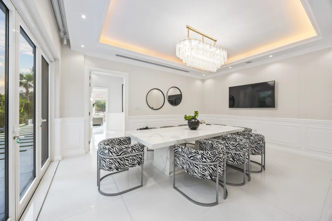 Bright modern dining room with a white marble table, zebra-print chairs, a crystal chandelier, and a wall-mounted TV.
