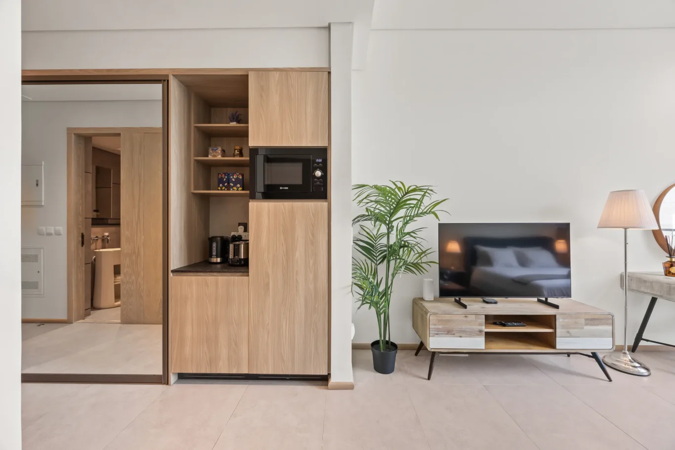 Open-plan living area with a built-in wooden cabinet housing a microwave, coffee station, and shelves beside a wall-mounted TV, plus a potted plant.