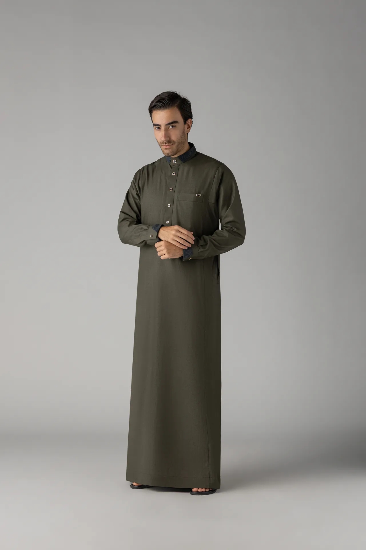Man in a long olive-green thobe stands against a plain gray background with hands clasped at waist level.