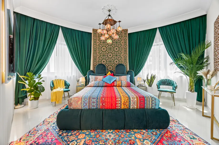 Luxurious bedroom with emerald drapes, a multicolored patterned quilt, and a teal upholstered bed with a decorative headboard and chandelier above.