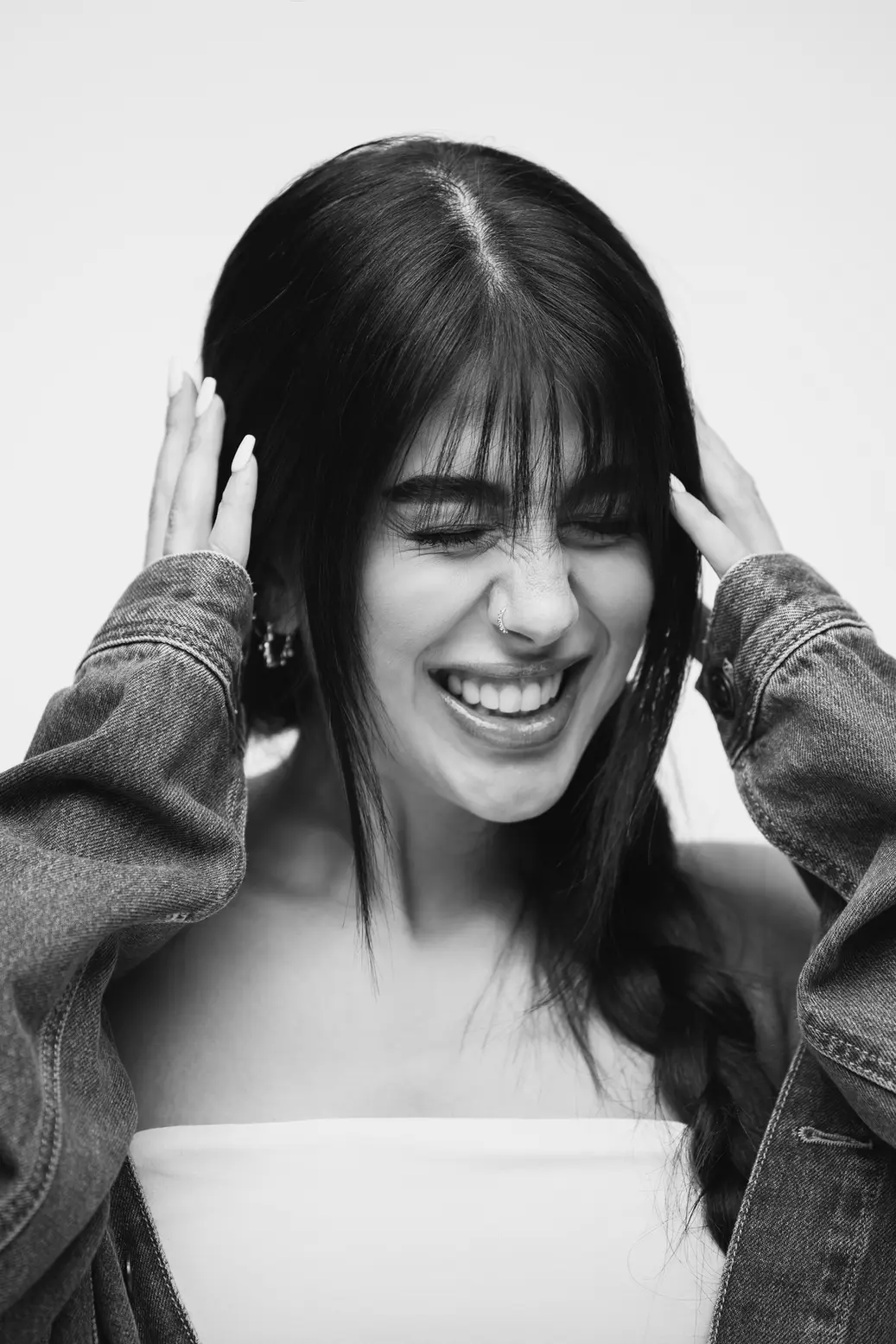 Smiling woman with long dark hair in a braid, hands near her head, wearing a denim jacket in a candid portrait (black and white).