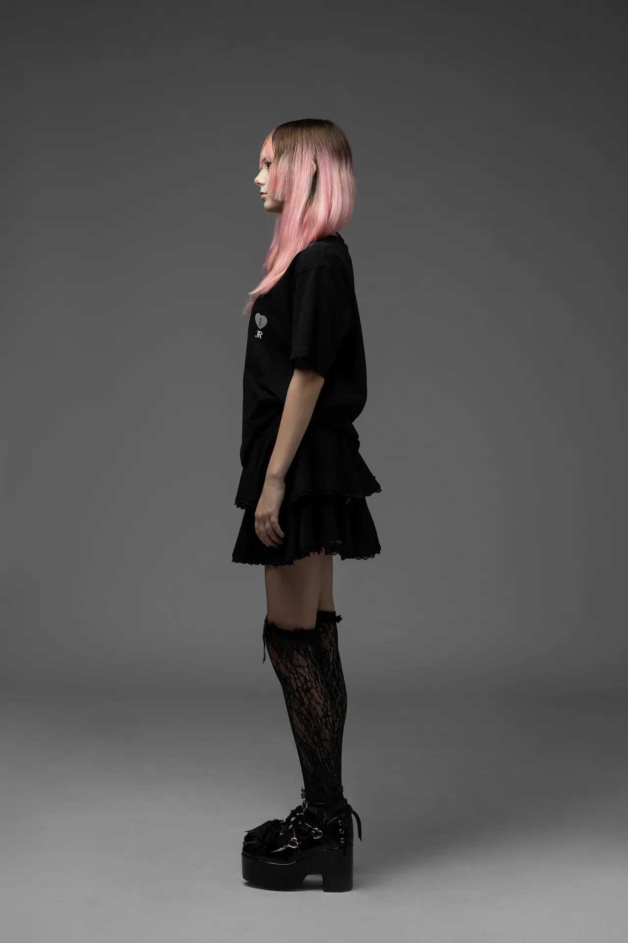 Side profile of a person with pink-tinted hair wearing a black layered dress, lace stockings, and platform shoes against a grey studio backdrop.