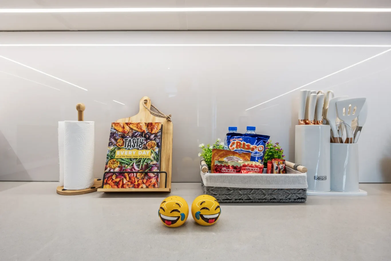 Modern kitchen counter with a paper towel roll on a wooden stand, a cutting board, and a rack of snack magazines beside a basket of chips; two emoji-face balls on the floor in front.