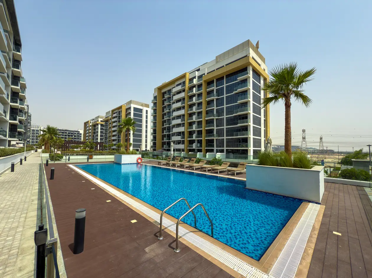 Outdoor swimming pool in a modern residential complex with lounge chairs and palm trees, multi-story buildings in the background under a clear blue sky like a resort.