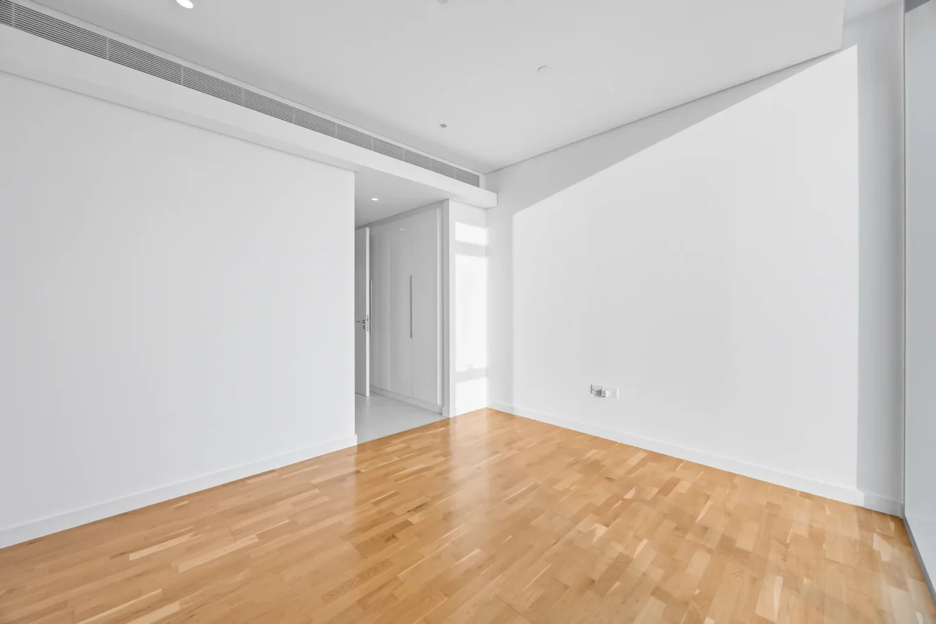 Empty contemporary room with light wood flooring, white walls, and a long ceiling vent; doorway to an adjacent space on the left.