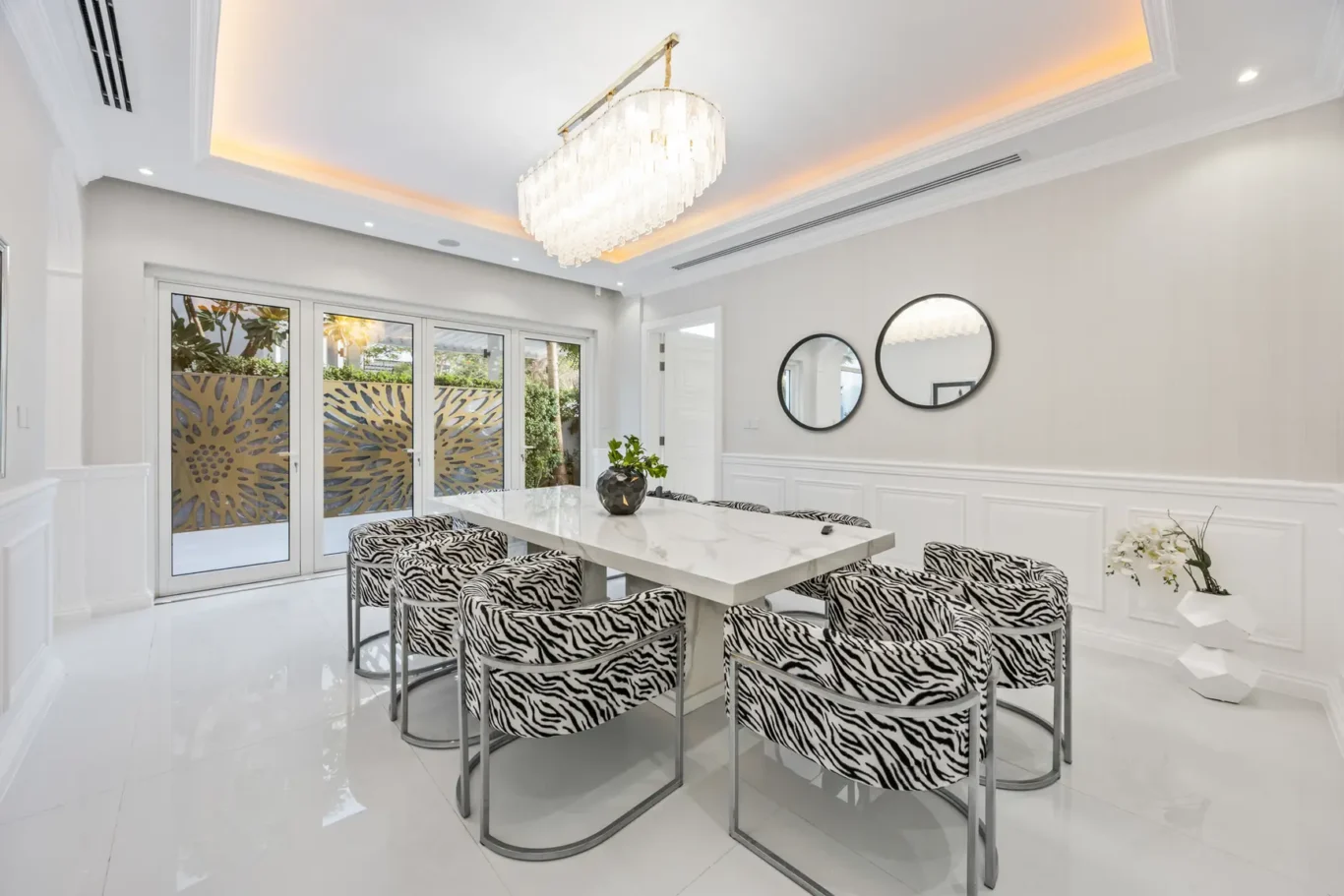 Modern dining room with zebra-print chairs around a white marble table and a crystal chandelier above. They sit on a glossy white floor beside frosted-glass doors with sunburst patterns.
