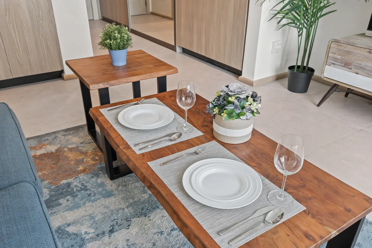 Wooden dining table set for two with gray placemats, white plates, silverware, and wine glasses; center plant arrangement appears as decor.