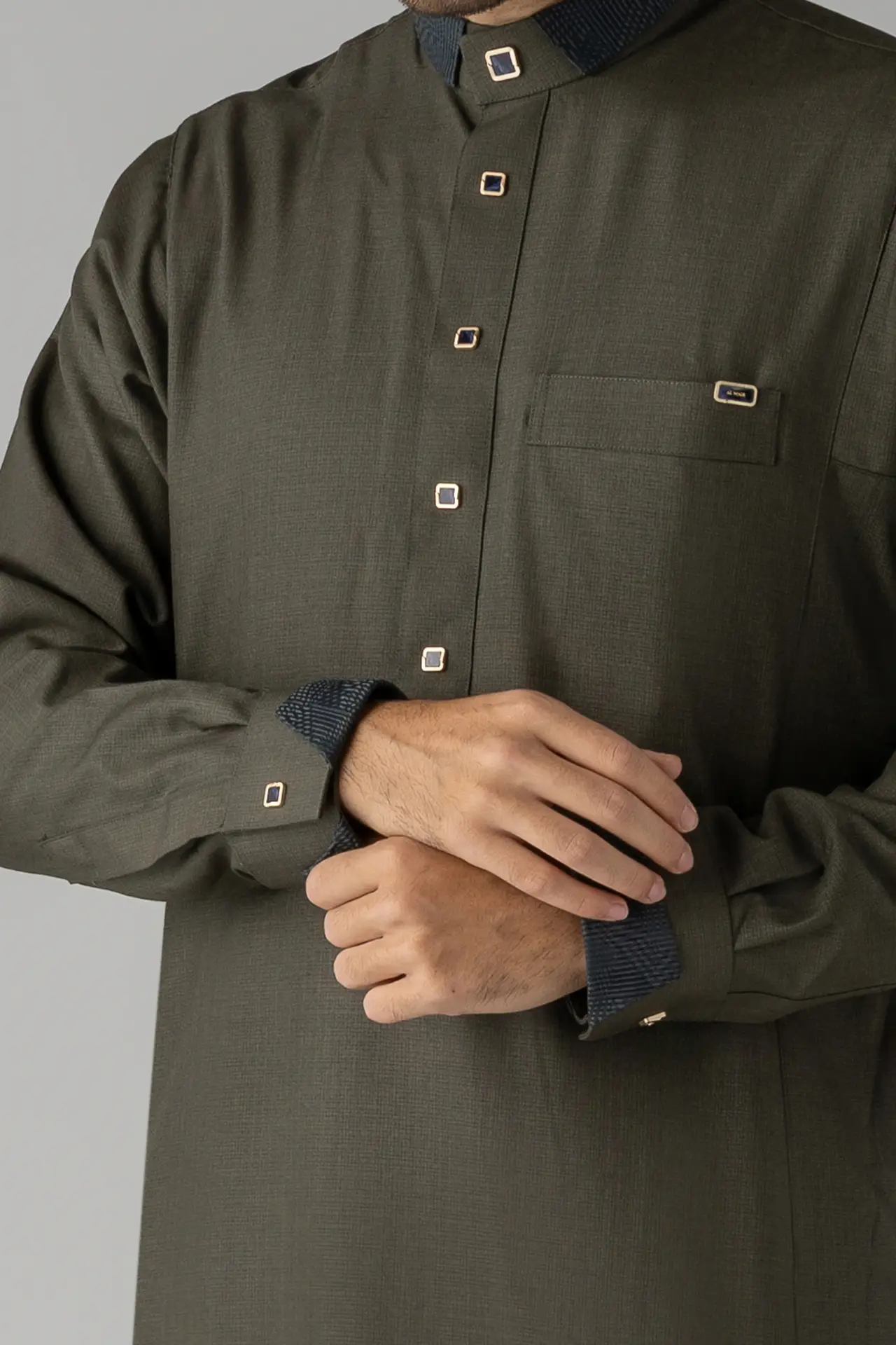 Close-up of a person wearing an olive green shirt with square gold buttons and blue cuffs, arms crossed.
