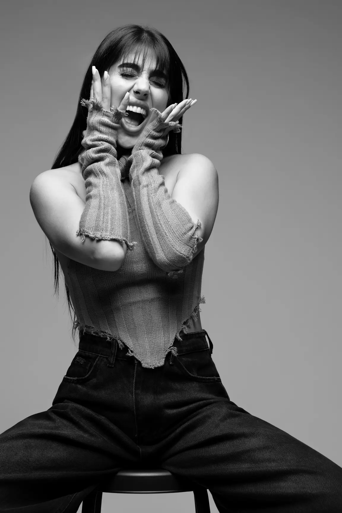 Woman laughing with hands on her cheeks, wearing a ripped knit top in a black-and-white studio photo.