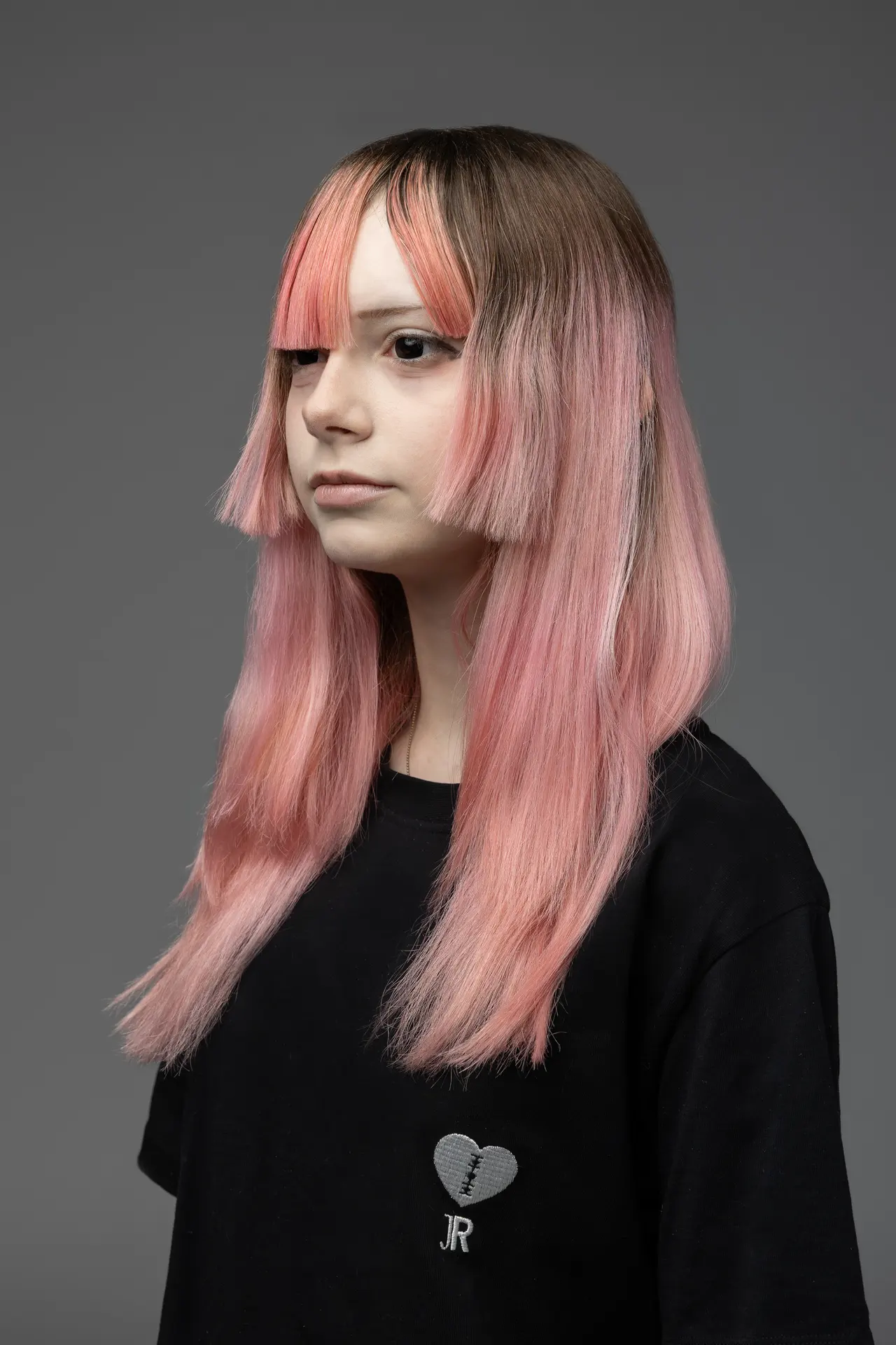 Side-profile portrait of a person with long pink ombré hair and blunt bangs, wearing a black shirt with a stitched heart graphic against a gray studio background.