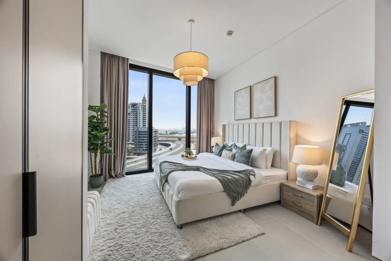 Bright hotel bedroom with a large upholstered headboard, two bedside lamps, and a floor-to-ceiling window with a city view.