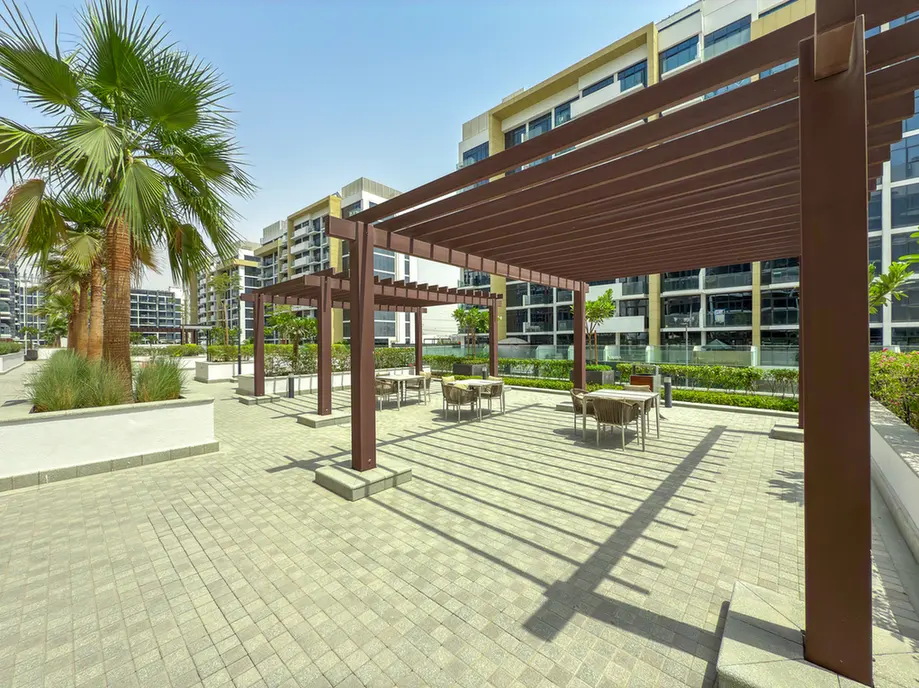 Open-air communal courtyard with brown pergolas, palm trees, and tables for outdoor dining or lounging in a modern residential complex.