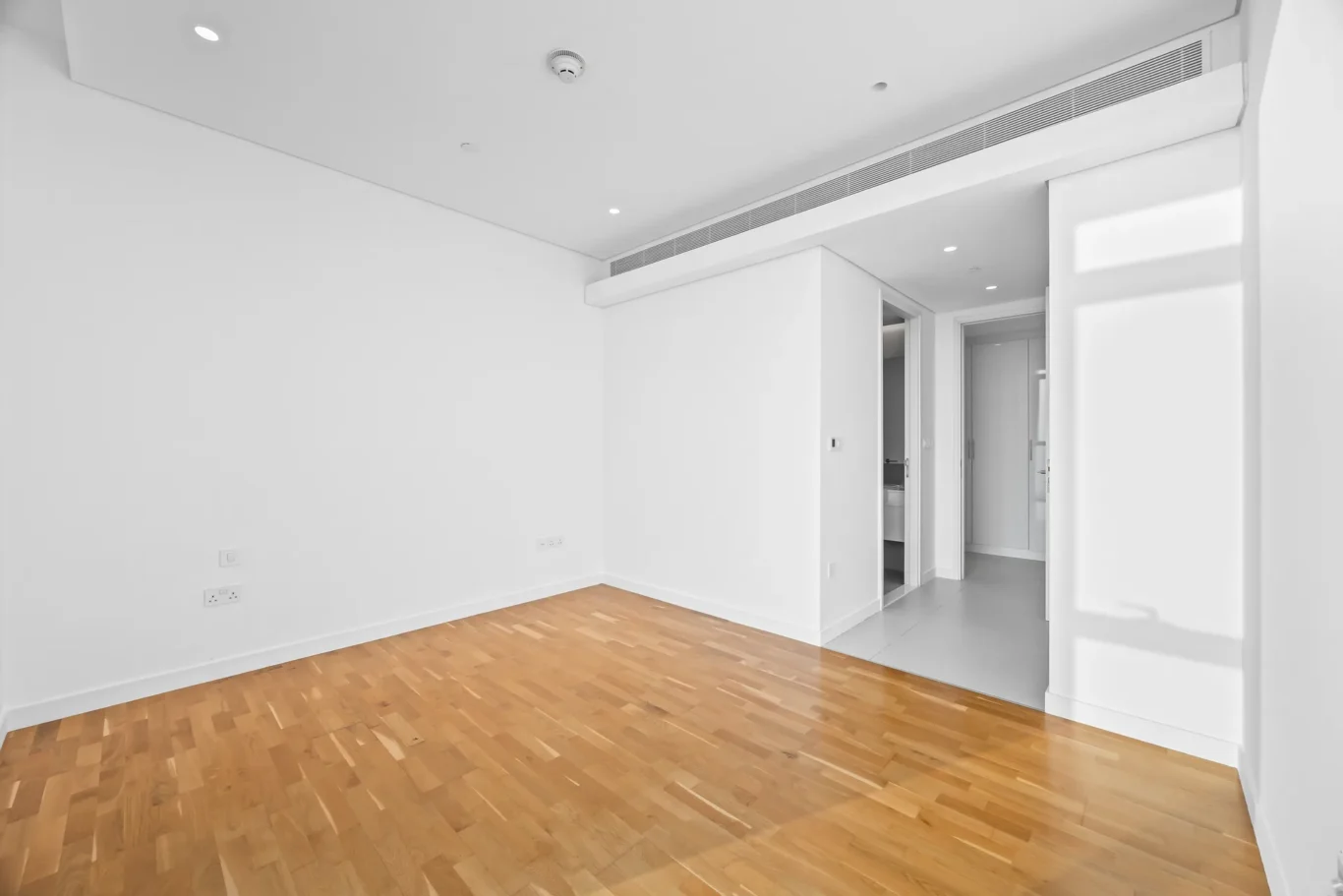 Bright empty room with white walls and light wood flooring, doorway to a hallway and visible ceiling vents/ducts.