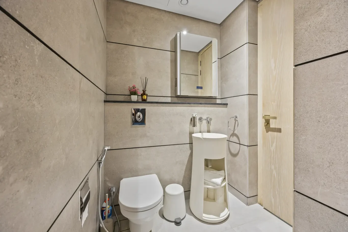 Small modern bathroom with beige tiled walls, wall-mounted toilet, curved open-shelving sink unit, and a large mirrored cabinet above. A wooden door is on the right.