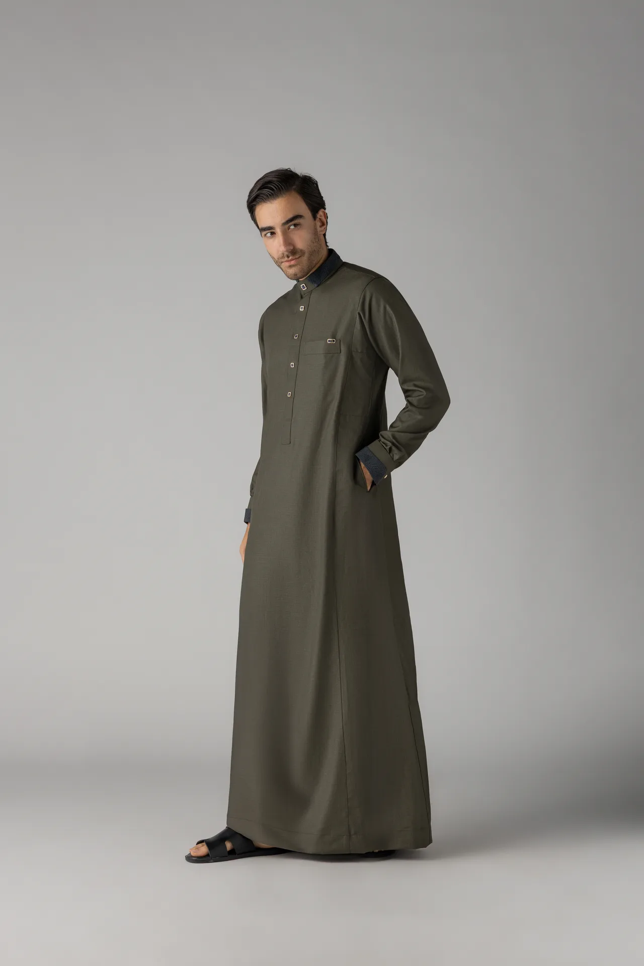 Young man in an olive-green traditional thobe, standing at a slight angle with hands in pockets against a neutral gray backdrop.