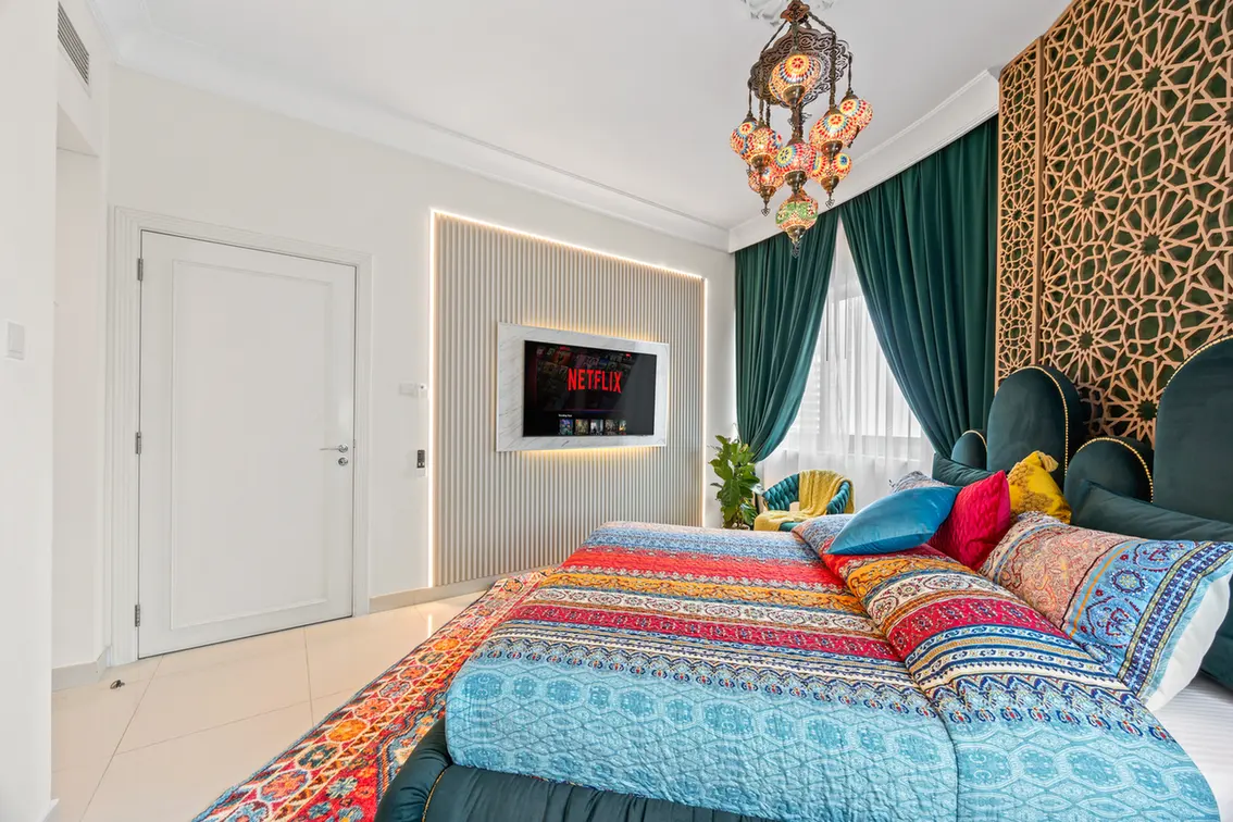 Luxurious bedroom with a large bed in vibrant patterned quilts, teal headboard, and emerald drapes framing a bright window.