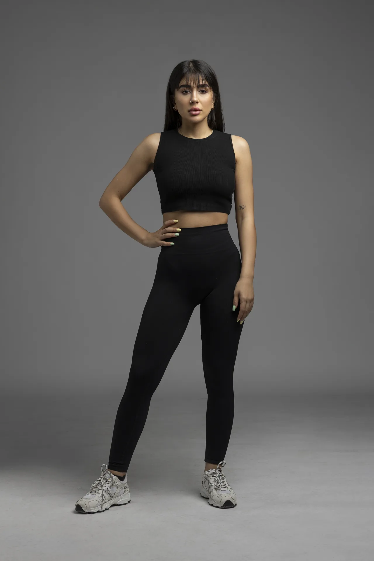 Woman in black athletic outfit standing with one hand on hip against a gray studio backdrop with a confident pose.