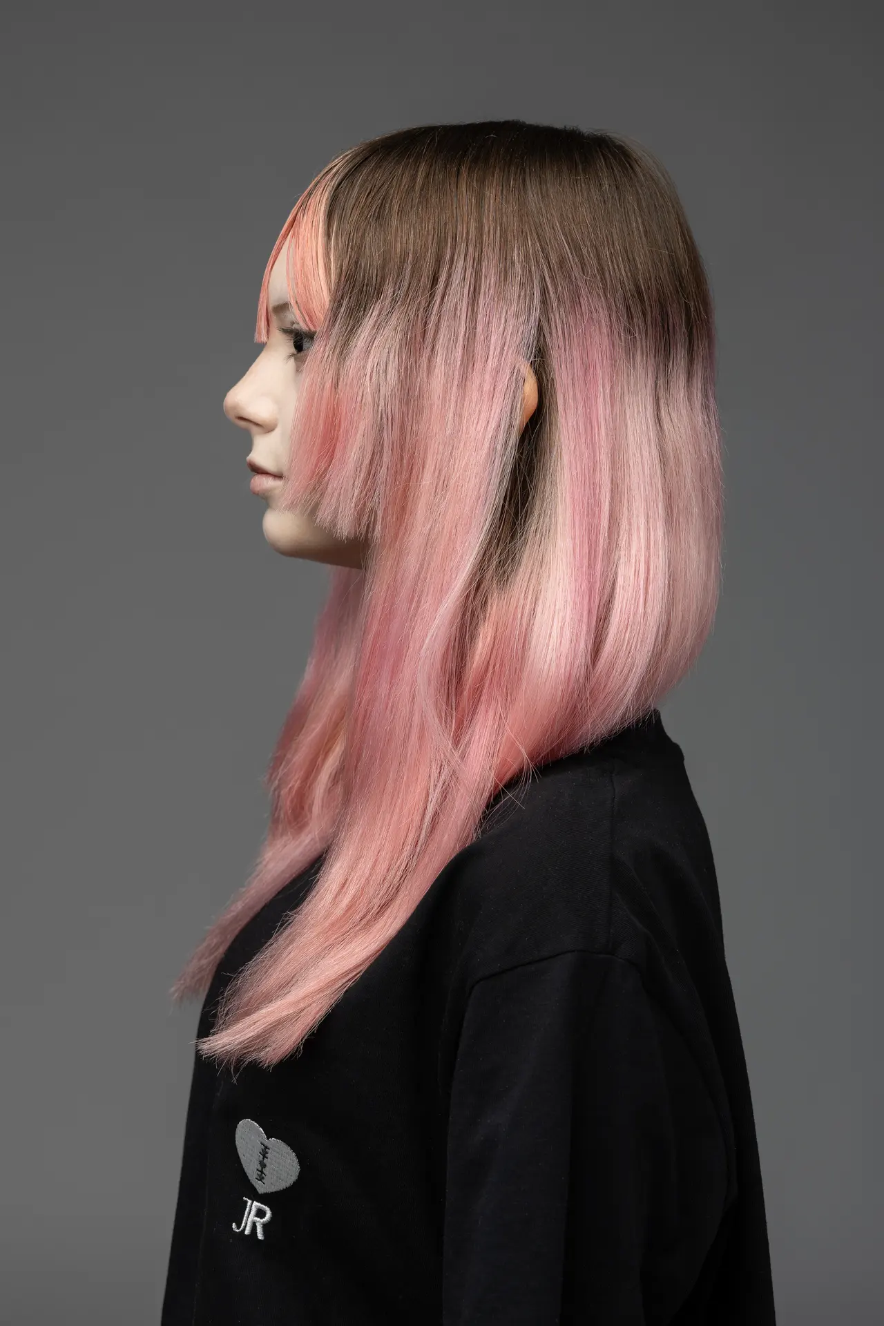 Side profile of a person with pink ombre hair fading from brown roots, wearing a black shirt against a gray backdrop.