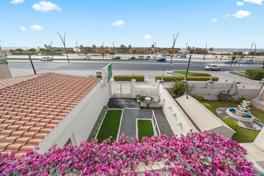View from a balcony of a courtyard with purple flowers in the foreground, tiled roof on the left, paved paths, a small fountain on the right, and a street with cars beyond a fence.