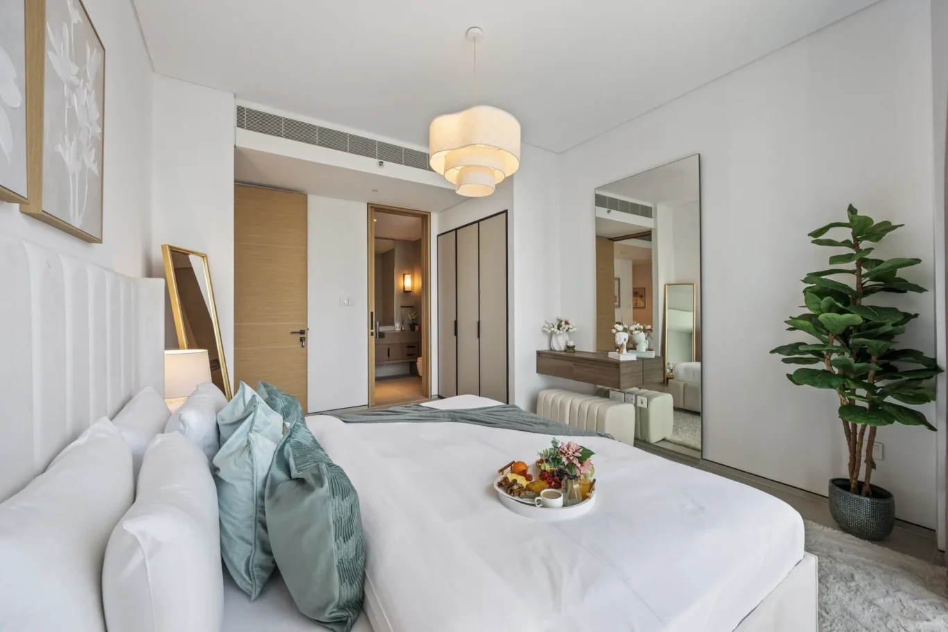 Hotel bedroom with a white upholstered bed and teal pillows; a fruit tray rests on the duvet in the foreground, mirrored by a large wall mirror opposite.