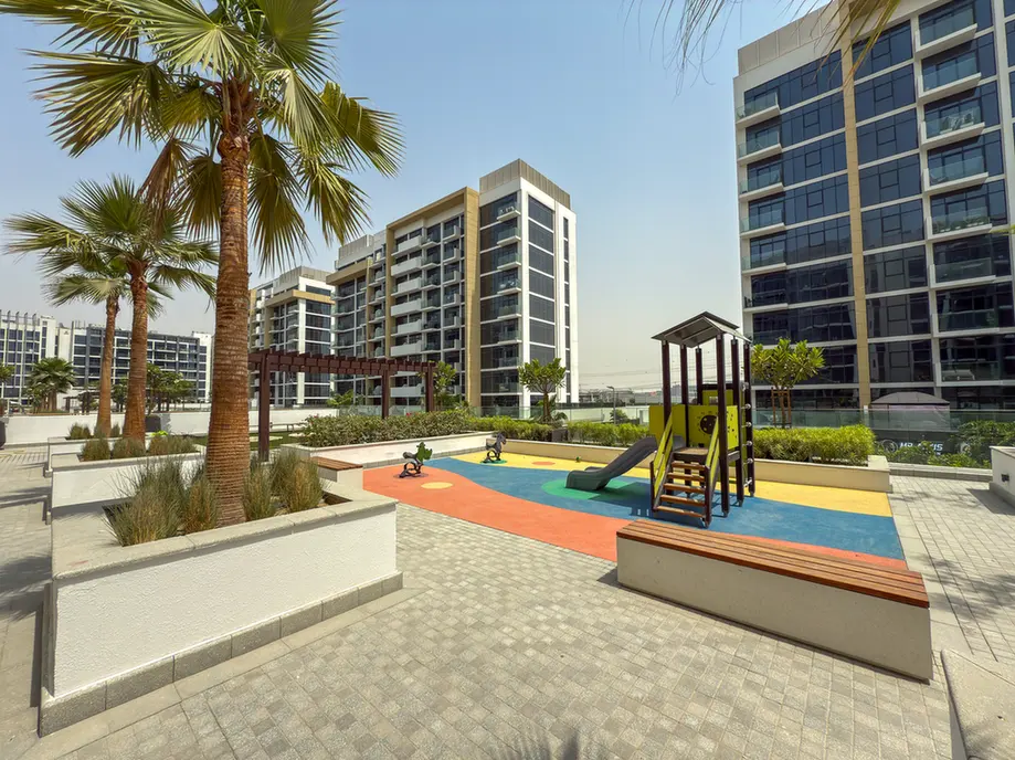 Children's playground with a multicolored rubber surface in a modern residential courtyard, surrounded by palm trees and tall buildings.