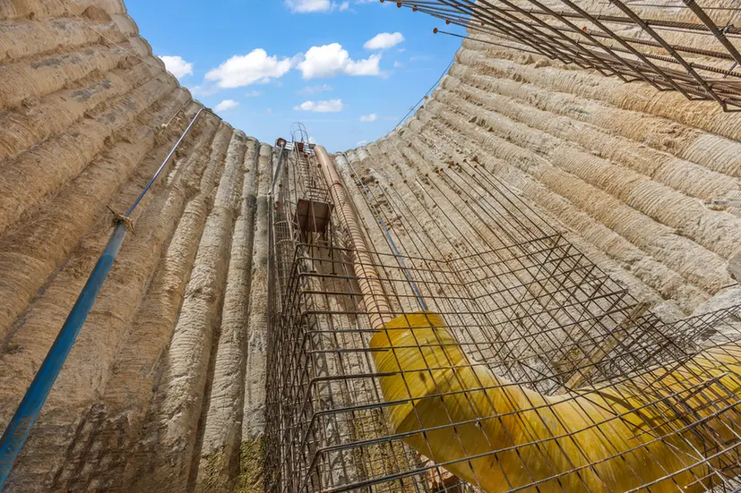Inside a large circular trench under construction with a grid of steel reinforcement bars; a worker in yellow gloves grips the bars against the bright blue sky.