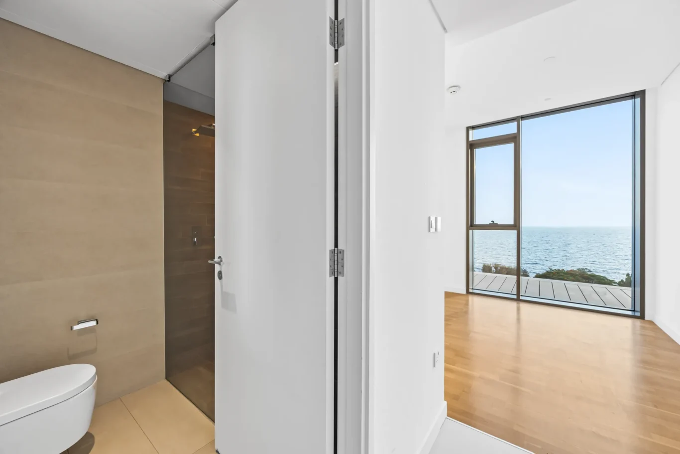 Modern bathroom with beige tiled walls and a white toilet, open white door reveals a bright room with large windows overlooking the sea and deck outside