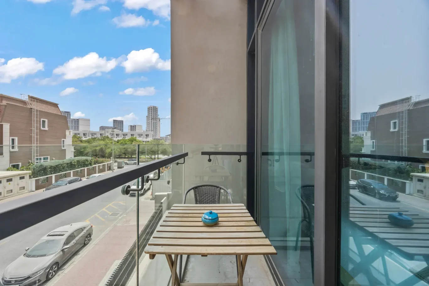 Balcony with a wooden folding table and a small blue teapot, glass railing, overlooking an urban street and blue sky.