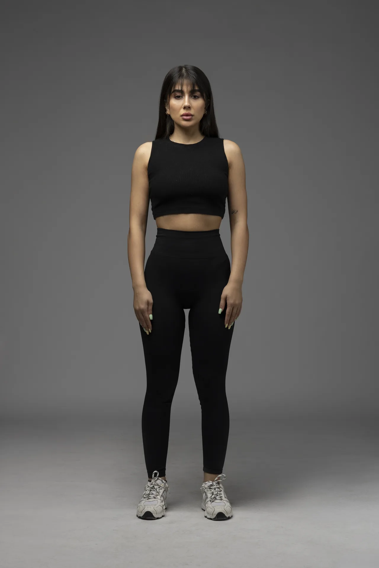 Front view of a woman in black athletic wear—a sleeveless crop top and high-waisted leggings—standing in a neutral gray studio background.