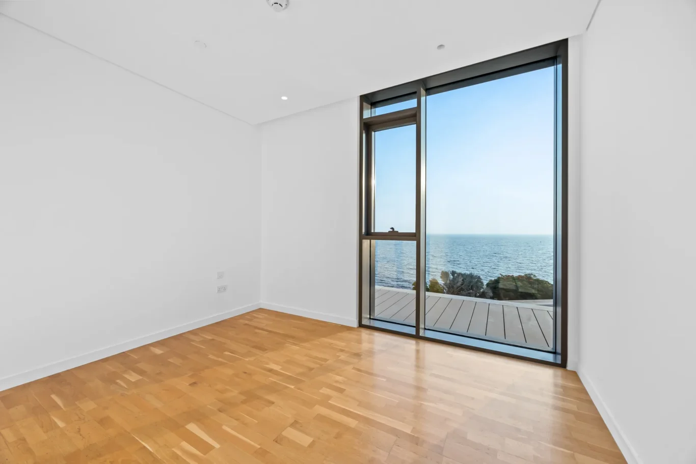 Empty white room with light wooden floor and a large glass door to a deck, offering an ocean view.
