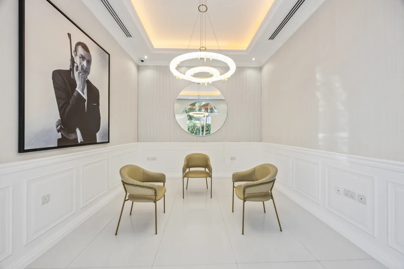 Modern white-walled waiting area with paneled wainscoting, beige chairs arranged in a circle, and a large black-and-white portrait of a man holding a gun on the left wall