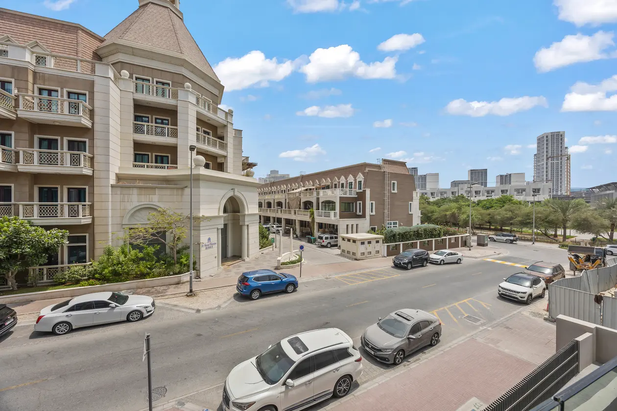 Wide street view of a modern urban neighborhood with beige stone buildings, parked cars, and a blue sky with scattered clouds.