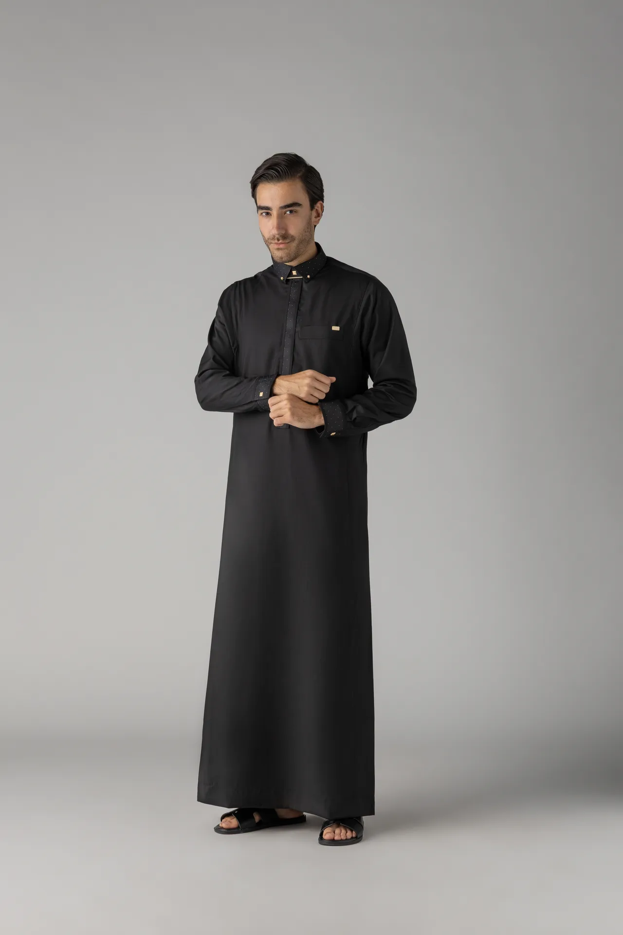 Man wearing a full-length black thobe and black sandals stands against a plain gray studio backdrop, looking at the camera with hands clasped.
