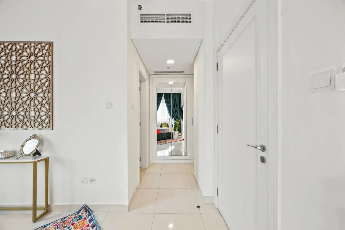 Bright white hallway with a decorative wooden panel on the left, a console table, and a doorway at the end showing a living room with teal curtains and a colorful rug in view through a mirror.