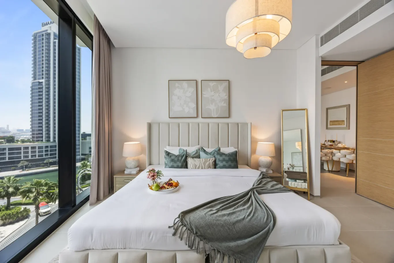Bright hotel bedroom with a large window showing a cityscape and water, a white tufted headboard bed, two lamps, and framed botanical art above the bed.