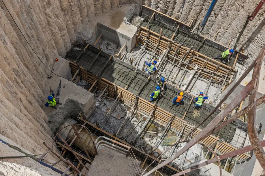 Construction workers in hard hats and high-visibility vests assemble concrete formwork and rebar in a deep excavation pit.