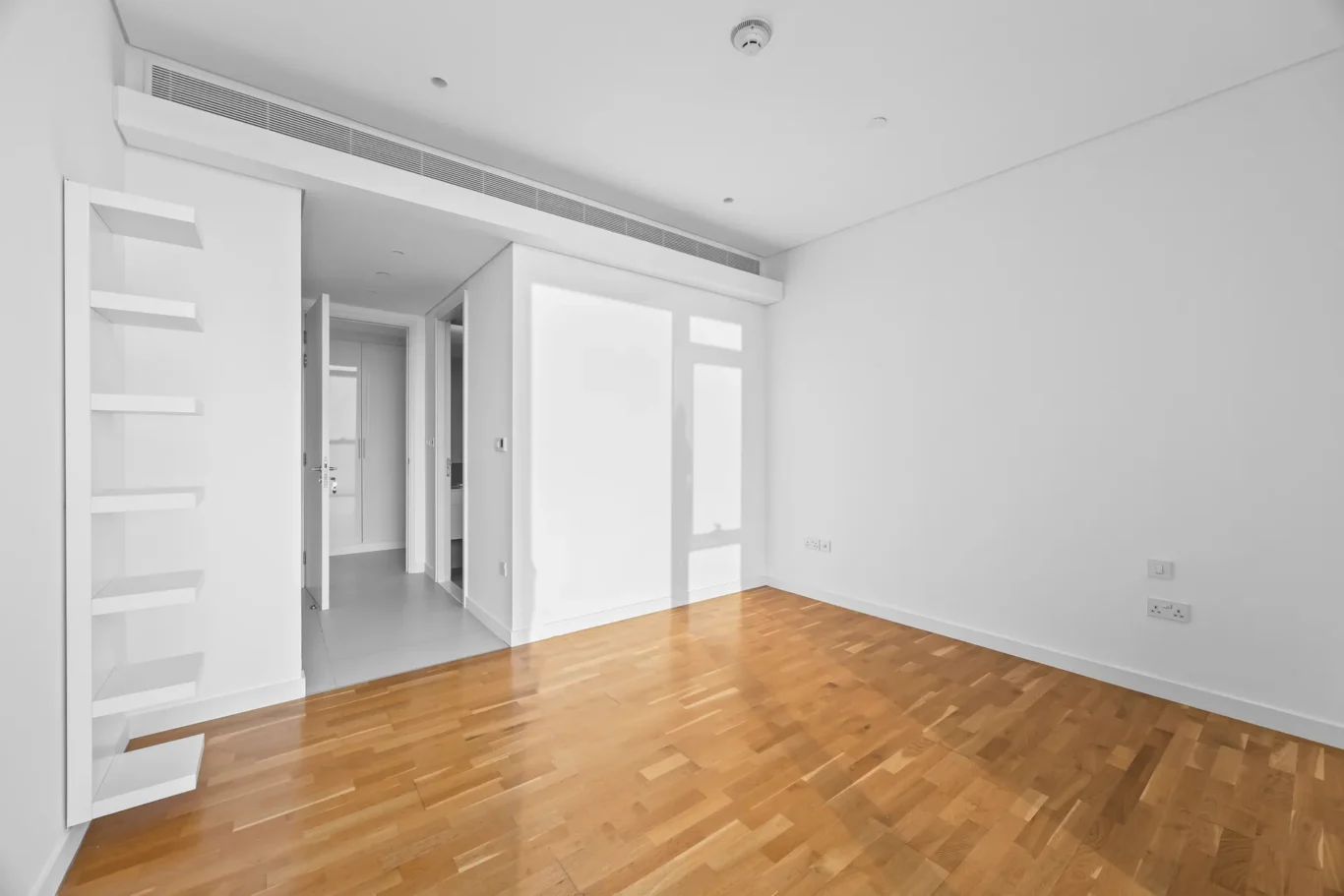 Empty white living room with warm wooden floor, built-in white shelves on the left, and a frosted glass door at the back.