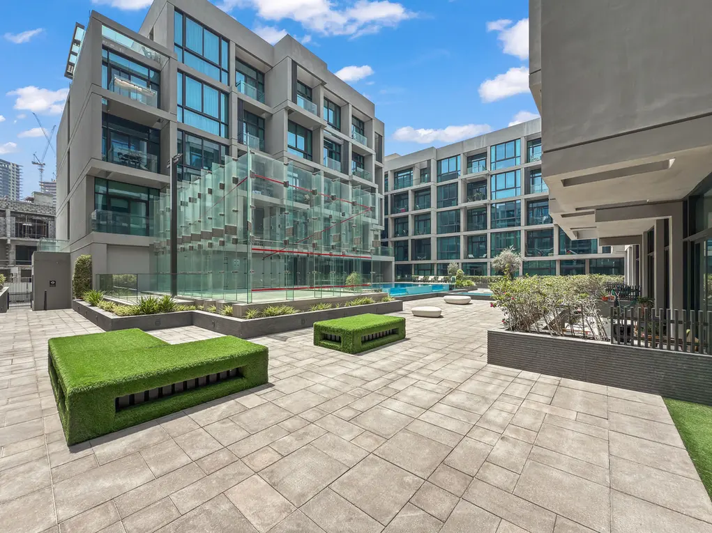 Modern courtyard of a contemporary building complex with glass balconies, a glass atrium, and a rectangular pool under a blue sky.