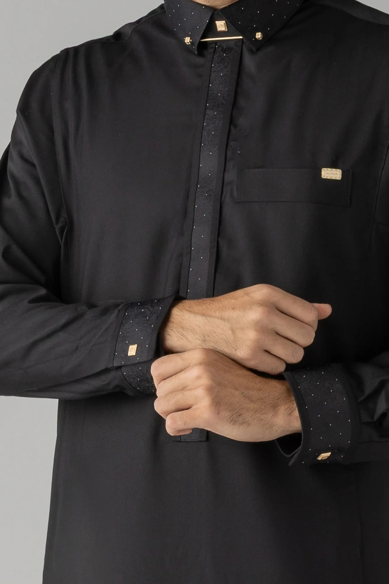 Close-up of a black dress shirt with small dotted pattern on the collar, placket, and cuffs, featuring gold pins and a pocket badge over the chest pocket nearby.