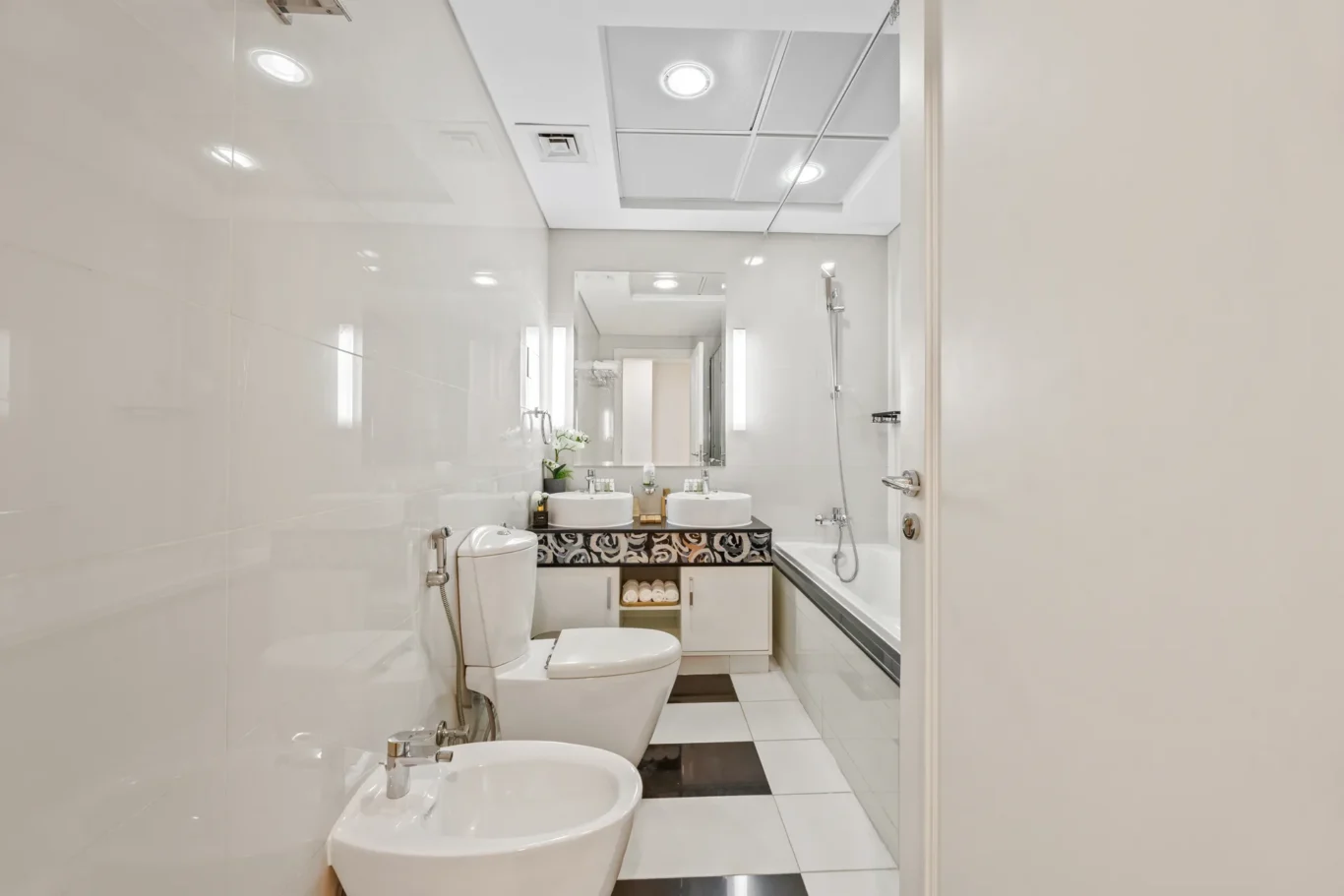 Bright, modern bathroom with two white sinks, a large mirror, and a bathtub along the right, viewed from the doorway.