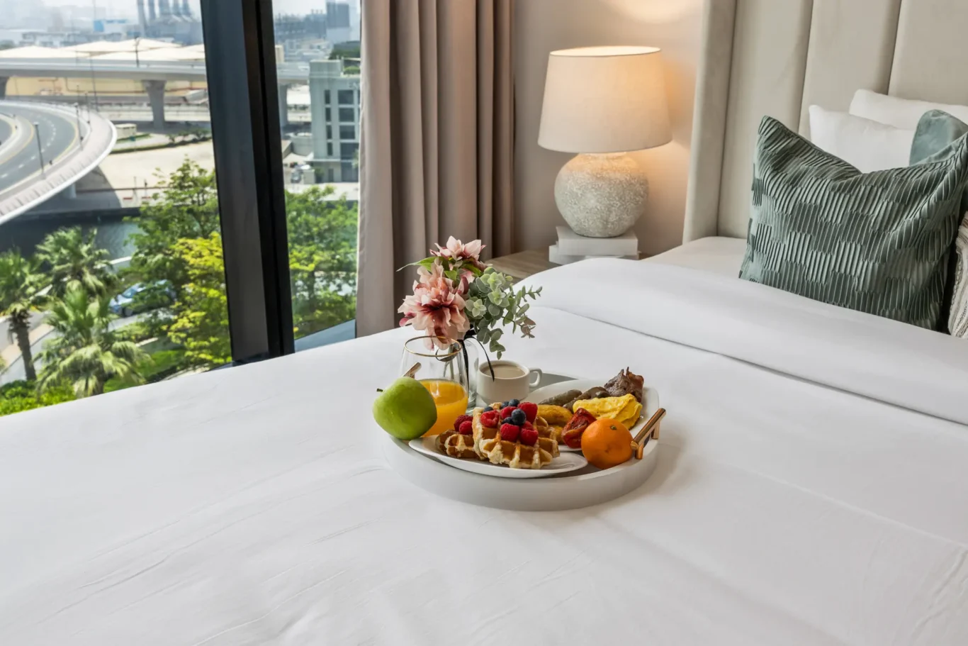 Breakfast tray on a white bedspread with fruit, pastries, and orange juice, by a window overlooking the city and greenery in a hotel room.