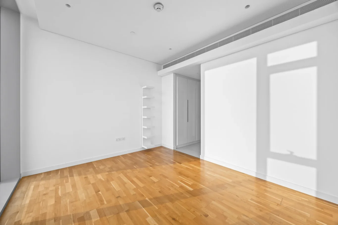 Bright empty white room with light wood floor, a tall white shelf unit against the left wall, and a built-in wardrobe nearby.