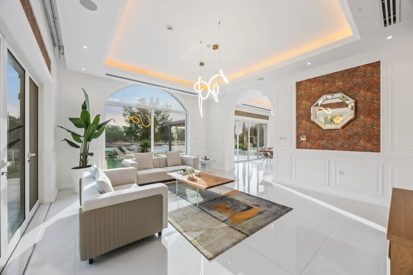 Bright modern living room with white glossy floors, beige sofa, wooden coffee table, and a large arched window overlooking an outdoor pool area; an orange textured wall with a mirrored octagonal frame stands to the right.