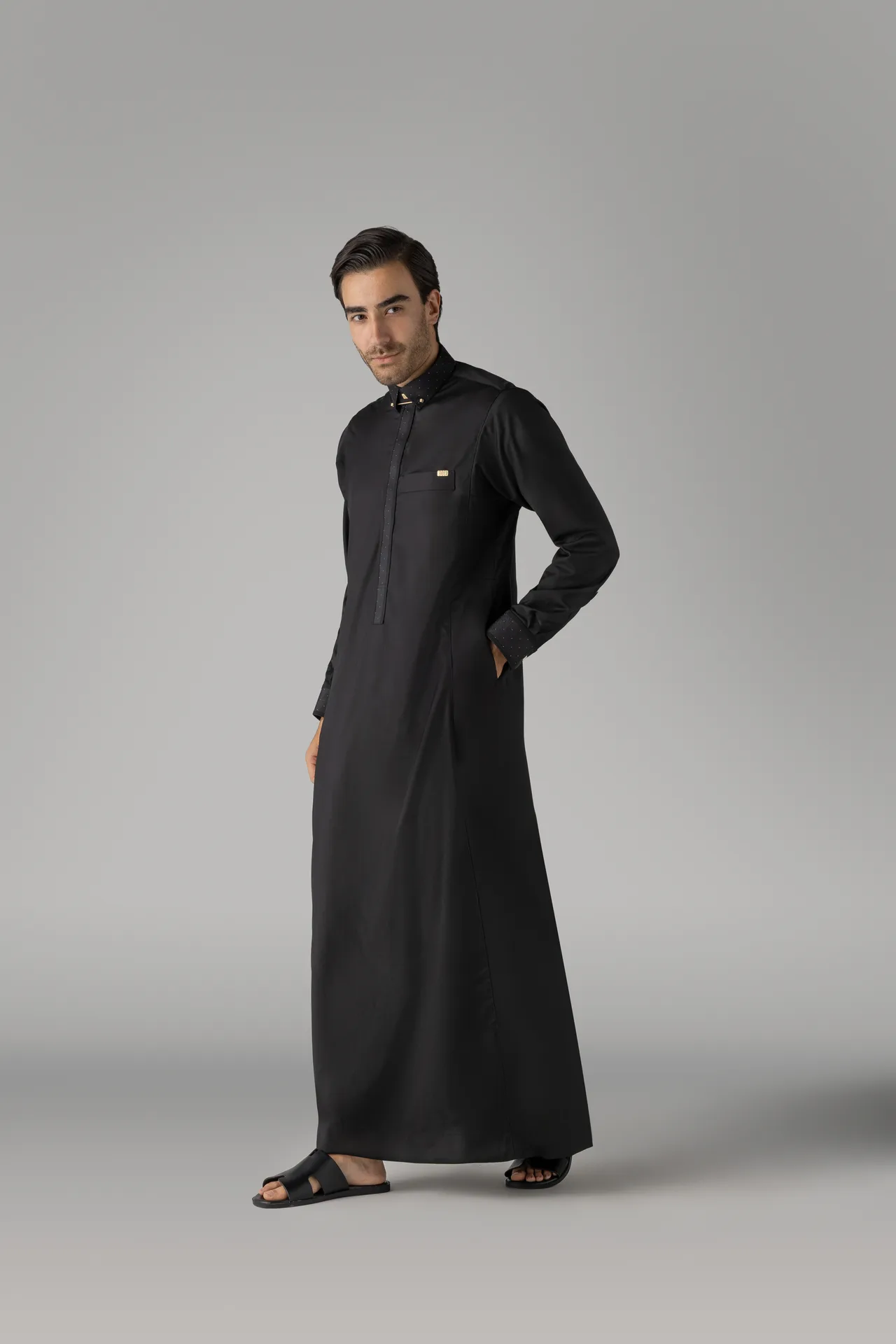 Man in a long black thobe with a subtle dotted collar, standing with one hand in pocket, wearing black sandals in a studio.