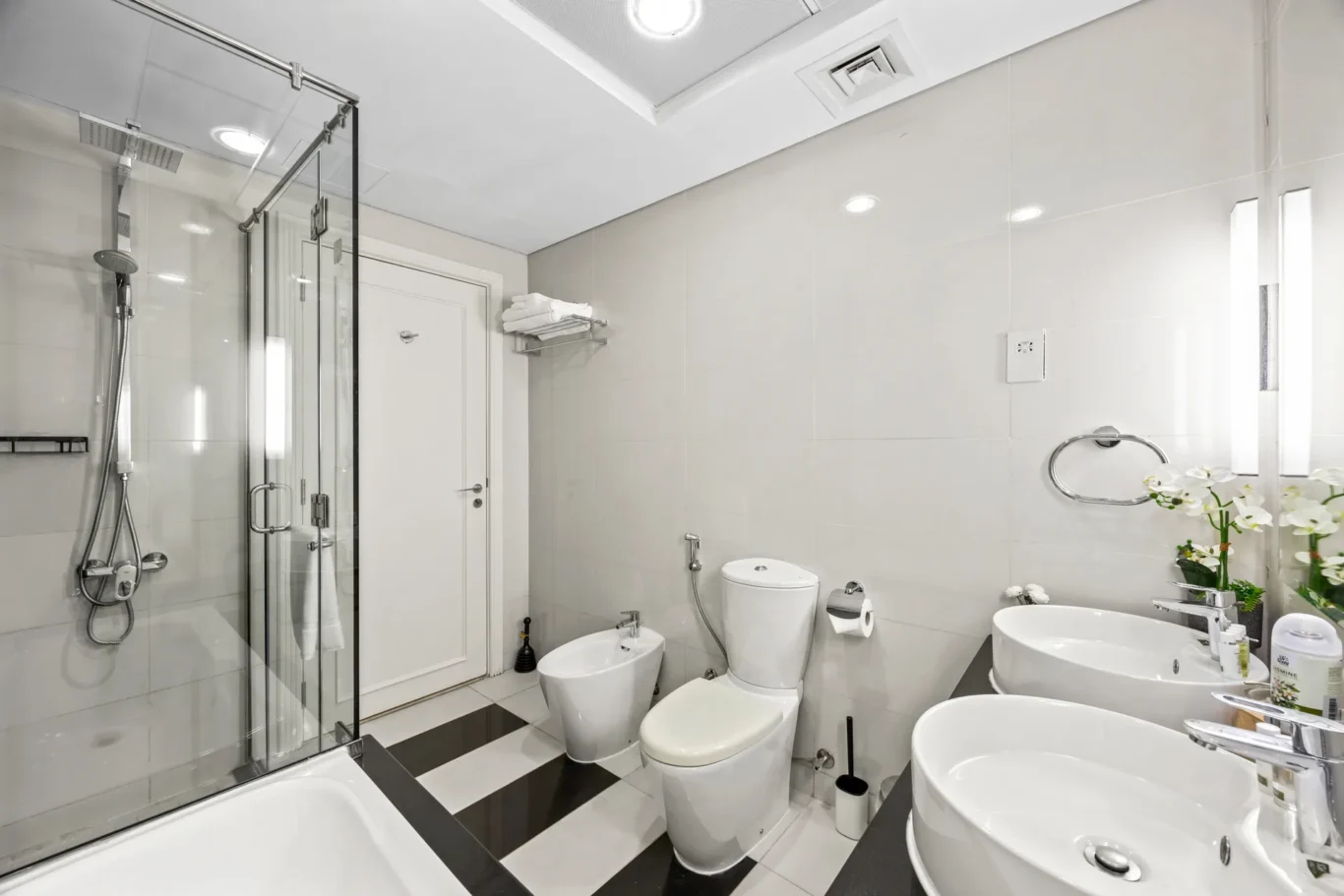 Modern white bathroom with glass shower enclosure, double sinks, toilet and bidet; elegant tiled walls and chrome fixtures.