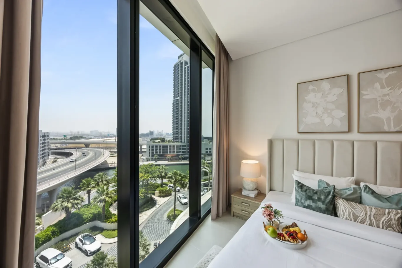 Hotel bedroom with a large window showing a cityscape and highway, a beige upholstered headboard, and a fruit tray on the bed.