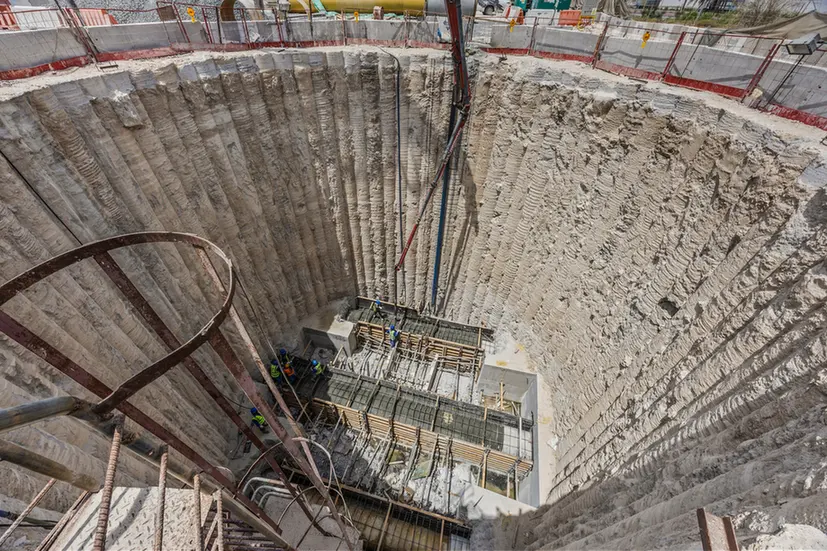 Deep circular excavation pit with stepped concrete walls, steel reinforcement, and workers assembling formwork at the bottom of a construction site.