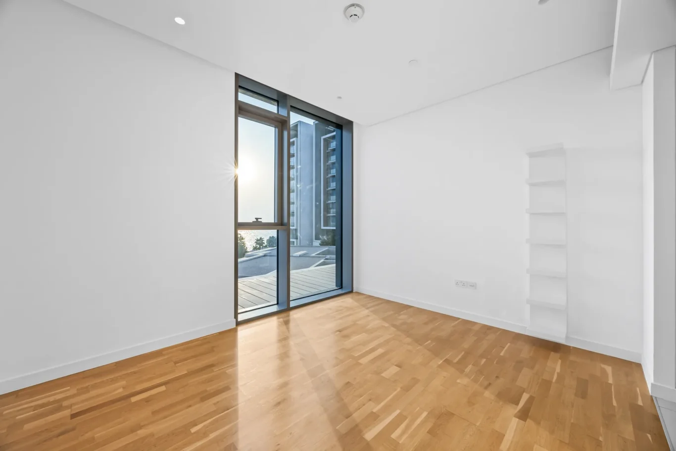Bright empty room with wood floor and large glass door to a balcony, overlooking a cityscape outside the window/door.