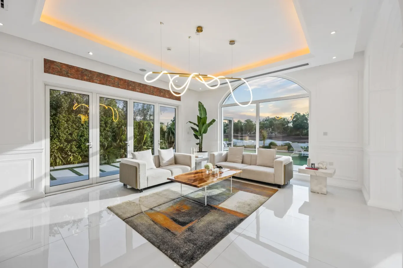 Bright modern living room with white walls, large glass doors, a sculptural pendant chandelier, beige sofas, and a wooden coffee table.
